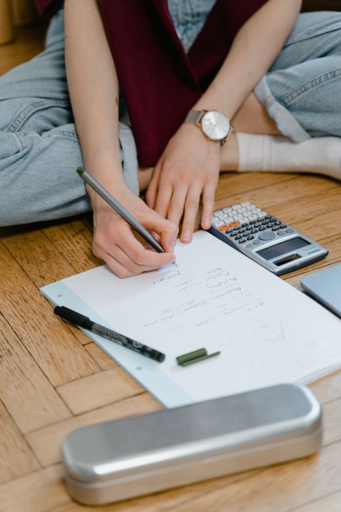A student writing notes on paper with calculator and pen on wooden floor.